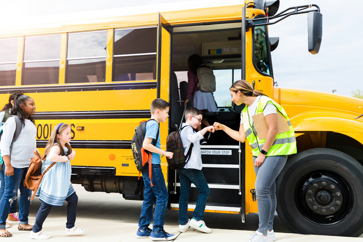 Cheerful female school bus driver or teacher gives students fist bumps as they board a school bus.